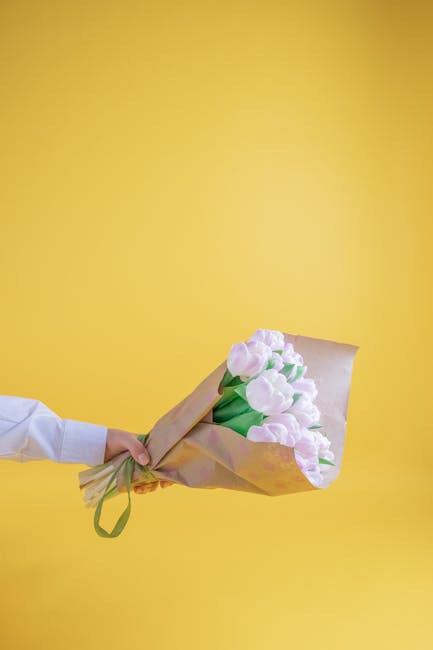 A person wearing a white shirt holds a bouquet of fresh, pale pink tulips wrapped in light brown kraft paper with a green ribbon against a bright yellow background. The tulips have smooth, rounded pet