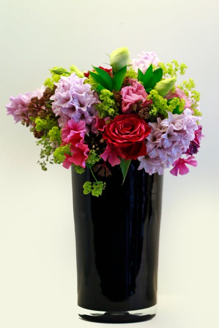 A tall, elegant black glass vase holds a vibrant bouquet of fresh flowers including a prominent red rose at the center, surrounded by soft pink and lavender hydrangeas, white lilies with tightly close