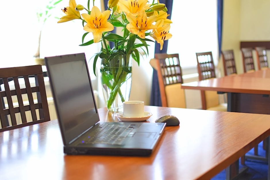 A clear glass vase filled with vibrant yellow lilies and green foliage sits on a polished wooden conference table, accompanied by a white coffee cup and saucer, a wireless mouse, and a silver laptop. 