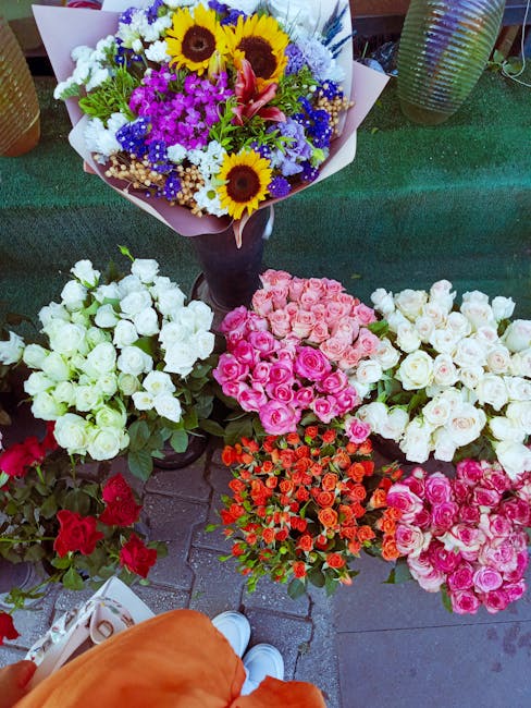 A vibrant display of freshly cut flower bouquets arranged outdoors on a tiled surface. The arrangement includes a large bouquet with sunflowers, purple statice, white chrysanthemums, pink lilies, and 