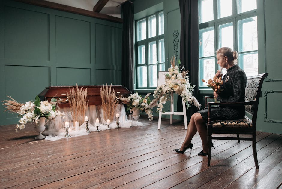 A woman dressed in black seated on a vintage-style armchair in a softly lit room with teal-green paneled walls and large windows. She is holding a small bouquet of flowers, possibly carnations and ros