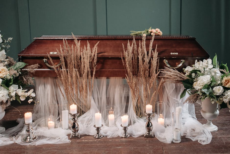 A polished wooden coffin placed on a wooden floor, surrounded by floral arrangements and candle decorations. The arrangements include white and cream roses, peonies, and other delicate blossoms set in