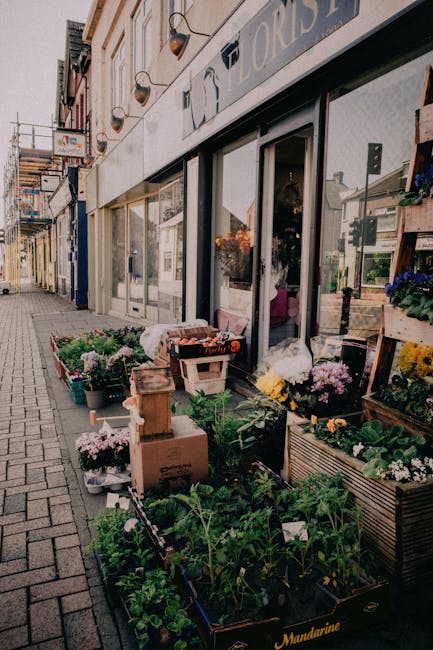 An exterior view of a flower shop on a city street, featuring a display of potted plants, flowering plants, and floral arrangements arranged on wooden crates and shelves outside the shop window. The f