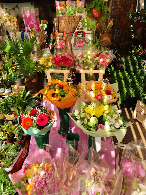 A display of fresh floral bouquets arranged on a pink cloth-covered table inside a florist shop, showcasing a variety of colorful flowers including yellow sunflowers, pink daisies, red roses, and whit