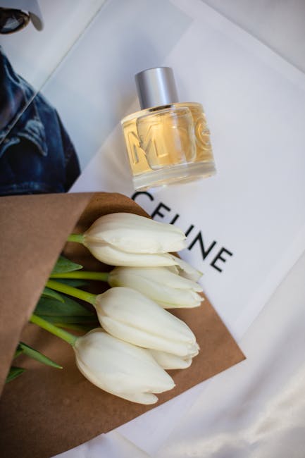 A bouquet of six white tulips with smooth, closed petals and green stems wrapped in brown paper, resting on a white surface. Nearby, there is a clear glass perfume bottle with a silver cap filled with