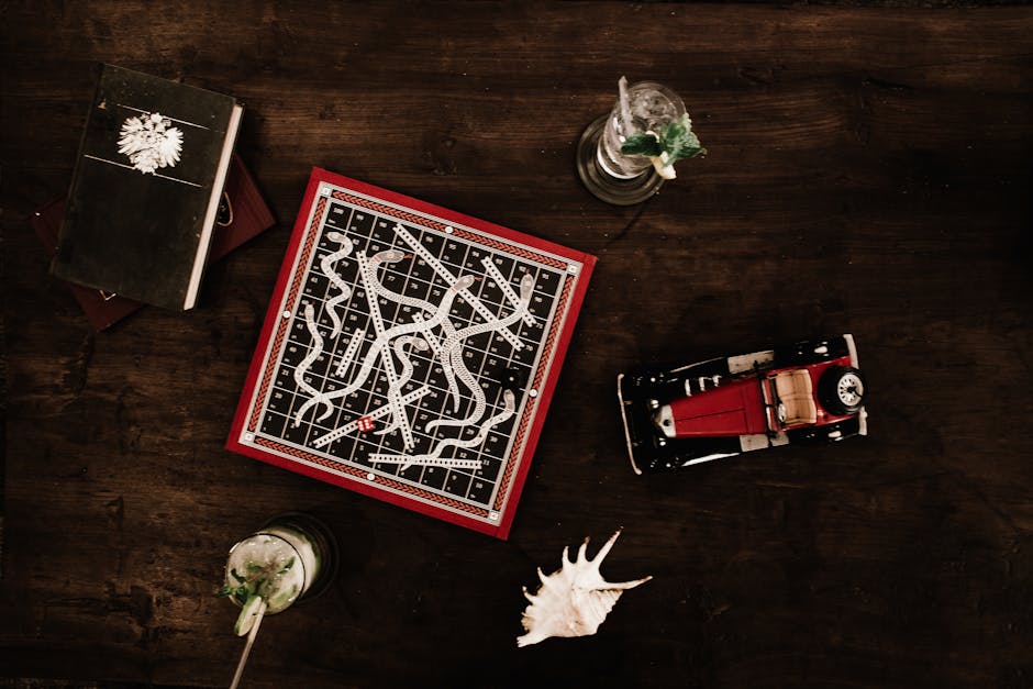 A top-down view of a dark wooden table displaying a vintage red and black toy car, a black book with a white wintery snowflake design, a square board game with a black and white winding path, and two 