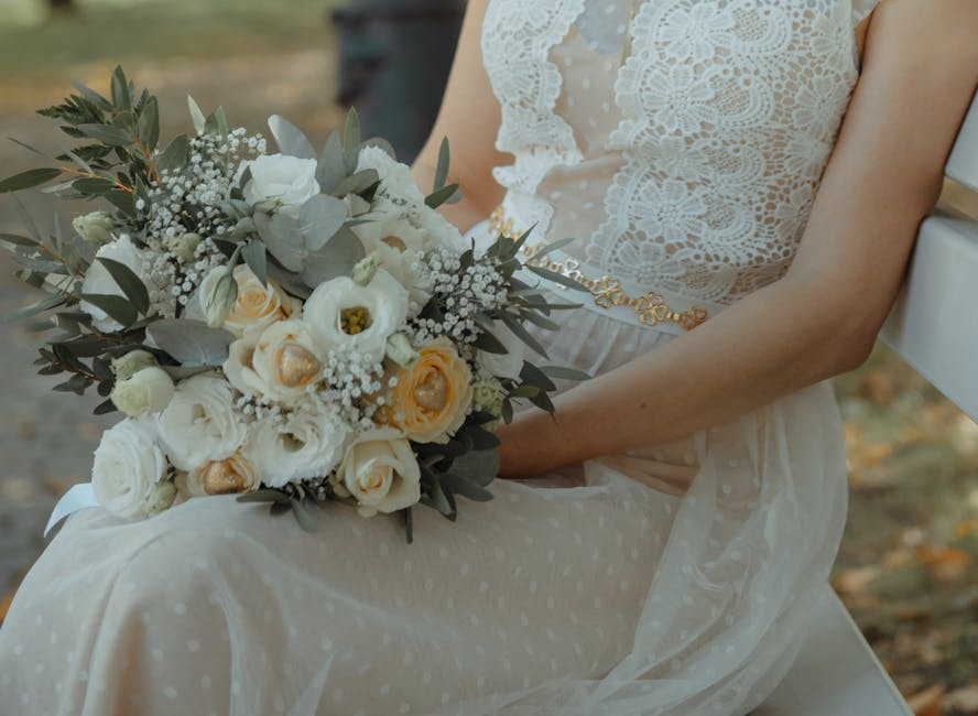 A close-up of a bride sitting on a white bench outdoors, holding a large floral bouquet comprised of white and cream roses, pale peach lisianthus, eucalyptus leaves, and delicate baby's breath. The bo