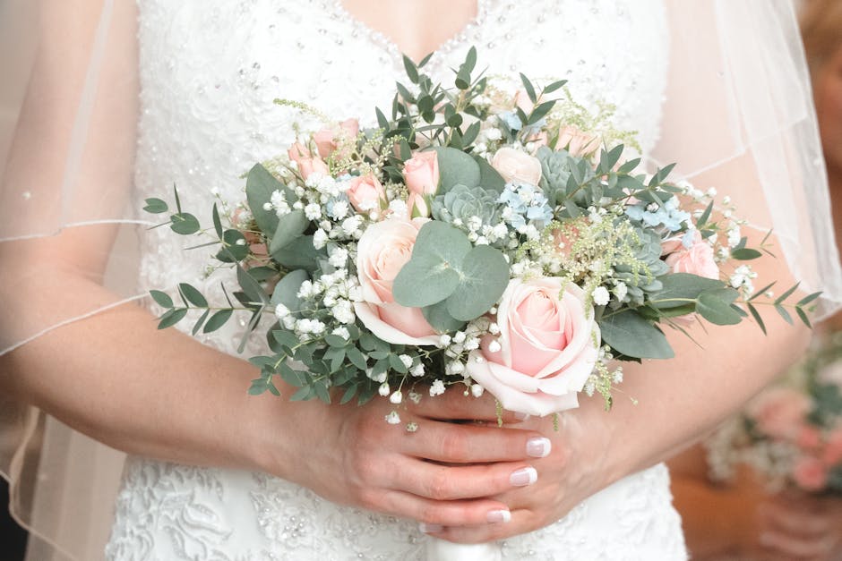 A bride holding a floral bouquet arrangement featuring pale pink roses, white baby's breath, eucalyptus leaves, and succulents, wrapped in neutral-toned paper with a satin ribbon. The arrangement is h