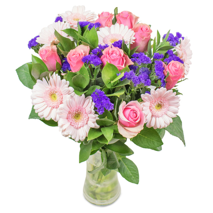 A masterfully arranged bouquet displayed in a slender clear glass vase, set against the soft hues typical of North London mornings. The focal point is a harmonious collection of 7-9 pink roses-some in tender, tightly furled buds, others just starting to spiral open. These blooms, each a marvel of classic form, are joined by 5-7 pale pink gerbera daisies, their wide, circular discs and feathered petals giving an appearance of airy fullness. Hints of almost-white at the daisies' centres create a luminous effect, further enhanced by scattered clusters of vibrant purple statice. The statice, with its crisp texture and wild aesthetic, introduces a striking contrast, reminiscent of the colourful markets along Harringay's busy high street. Deep, glossy green leaves weave around the base, abundant and fresh, their cool fragrance recalling early strolls through Fairland Park. All stems sit visible and clean in the water-filled vase, adding an organic transparency that signals freshness and care. This arrangement-featuring roses, gerbera daisies, and rich purple fillers-offers a thoughtful choice for birthdays, anniversaries, or as an elegant everyday accent for a Harringay home or office, delivering a visual message of celebration and affection unique to the area.
